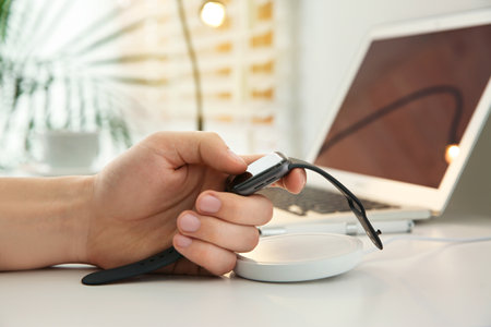 Man taking smartwatch from wireless charger at white table, closeup. Modern workplace accessories