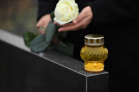 Woman holding white rose near black granite tombstone with candle outdoors, closeup. Funeral ceremonyの写真素材