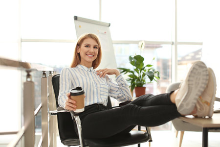 Young businesswoman with cup of coffee relaxing at table in office during breakの写真素材