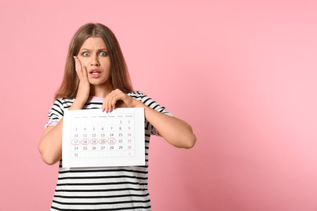 Emotional young woman holding calendar with marked menstrual cycle days on pink background. Space for textの写真素材