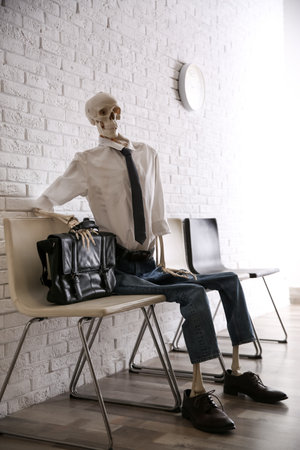 Human skeleton in office wear sitting on chair near brick wall indoorsの写真素材