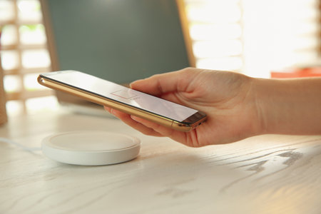 Woman putting mobile phone onto wireless charger at white wooden table, closeup. Modern workplace accessoriesの写真素材