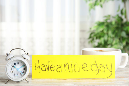 Delicious morning coffee, alarm clock and card with HAVE A NICE DAY wish on white wooden table indoorsの写真素材