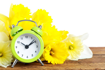Green alarm clock and spring flowers on wooden table against white background. time changeの写真素材