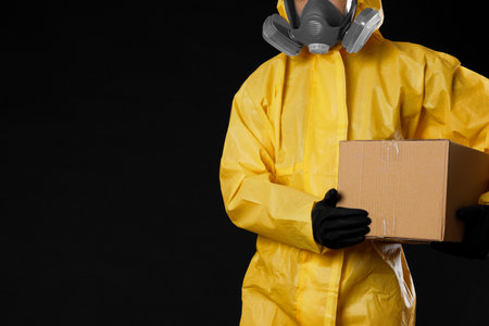 Man with cardboard box wearing chemical protective suit and space for text on black background, closeup. Prevention of virus spreadの写真素材