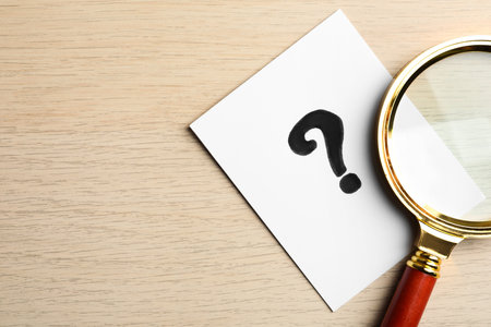 Paper card with question mark and magnifier glass on wooden table, top view. Space for text