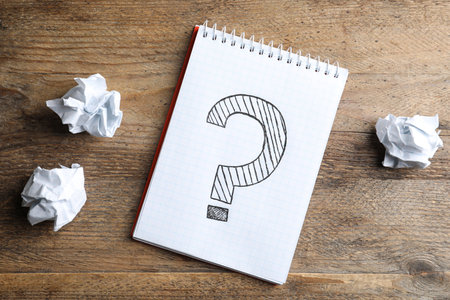 Notebook with question mark and crumpled paper on wooden table, flat lay