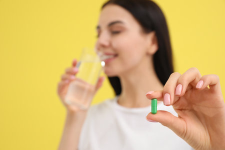 Young woman with glass of water and vitamin capsule against yellow background, focus on handの写真素材