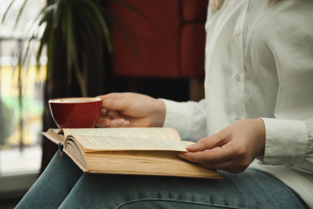 Woman with cup of coffee reading book indoors, closeupの写真素材