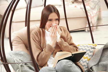 Sick young woman sneezing while reading book at home. Influenza virusの写真素材