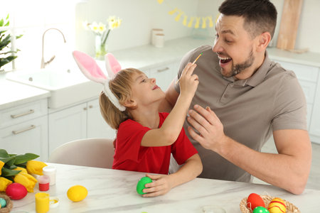 Happy son with bunny ears headband and his father having fun while painting Easter egg at table in...