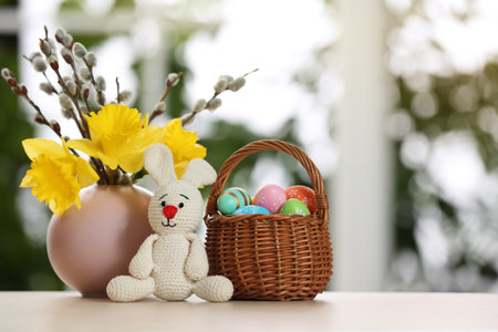 Festive composition with Easter eggs in wicker basket and bunny on table against blurred window,...