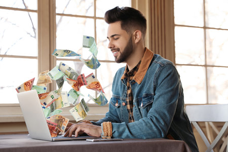 Man with modern laptop and flying euro banknotes at table indoors. people make money online