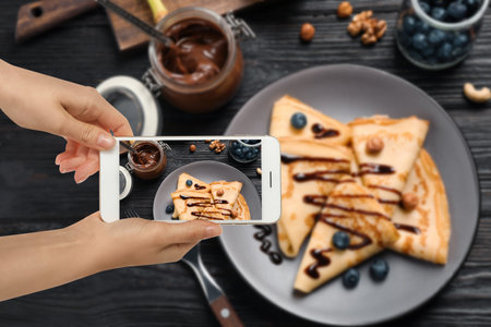 Food blogger taking picture of delicious thin pancakes on wooden table, closeup