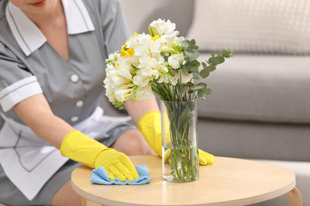 Young chambermaid wiping dust from wooden table in hotel room, closeup