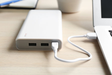 Charging modern laptop with power bank on wooden table, closeup