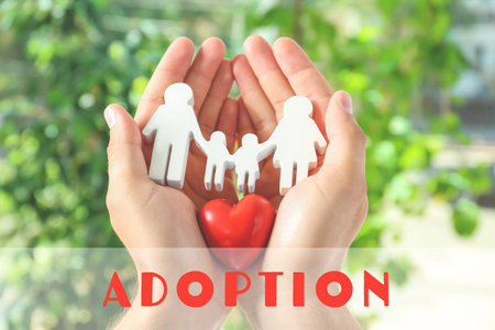 Young man holding wooden family figure and red heart in his hands against blurred background, closeup. child adoption conceptの写真素材