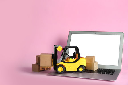 Laptop, forklift model and cardboard boxes on pink background. Courier serviceの写真素材