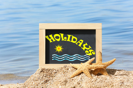 Little blackboard with word HOLIDAYS and sea stars on sand near seaの写真素材