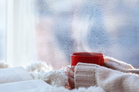 Woman with cup of hot drink near window on rainy day, closeupの写真素材