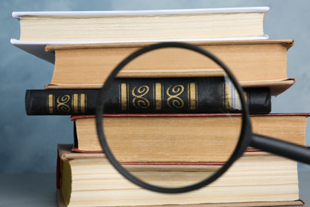 Looking through magnifying glass at stack of vintage books on gray table, closeup. Search concept