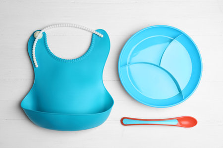 Colorful plastic dishware and silicone bib on white wooden table, flat lay. Serving baby foodの写真素材