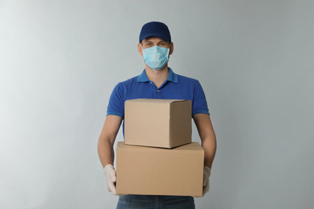 Courier in protective mask and gloves holding cardboard boxes on light background.の写真素材