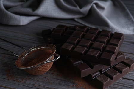 Tasty dark chocolate bars and sieve with cocoa powder on wooden table