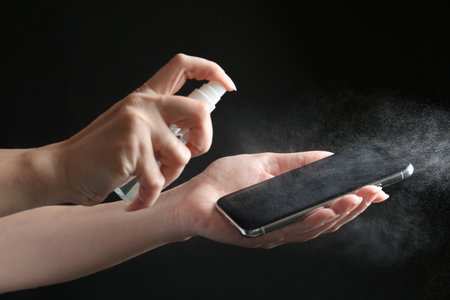 Woman spraying antiseptic onto smartphone against black background, closeup
