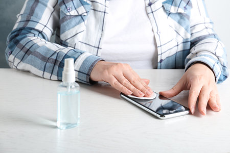Woman cleaning mobile phone with cotton pad and antiseptic at white table, closeup