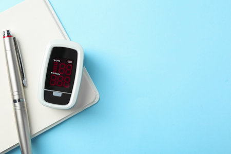 Modern fingertip pulse oximeter, pen and open notebook on light blue background, flat lay. Space for textの写真素材