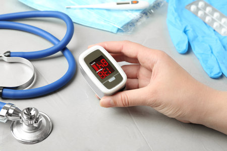 Woman holding fingertip pulse oximeter near medical items at light gray stone table, closeup