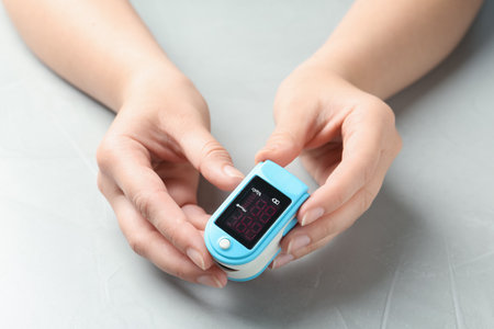 Woman holding fingertip pulse oximeter at light gray stone table, closeup