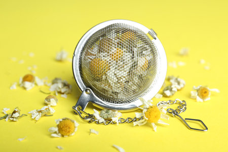 Dry chamomile flowers in infuser on yellow background, closeup