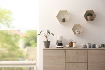 Modern coffeemaker and cups on commode near light wall indoors
