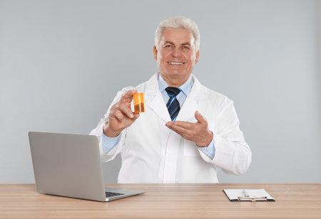 Professional pharmacist with pills and laptop at table against light gray backgroundの写真素材