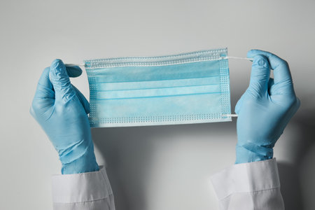 Doctor in latex gloves holding disposable face mask on light background, closeup.