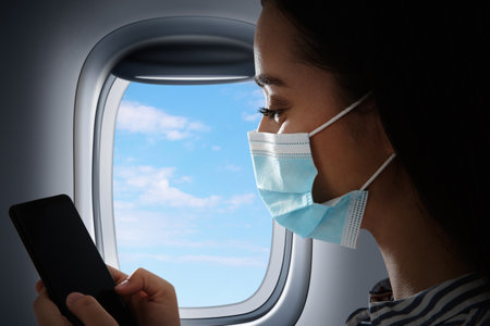 Traveling by airplane during  virus pandemic. Woman with face mask and phone near portholeの写真素材