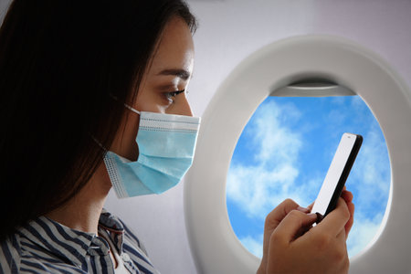 Traveling by airplane during   pandemic. Woman with face mask and phone near portholeの写真素材