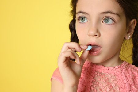 Little girl taking vitamin pill on yellow background, closeup. Space for textの写真素材
