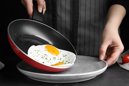 Woman putting tasty cooked eggs onto plate from frying pan at black table, closeupの写真素材