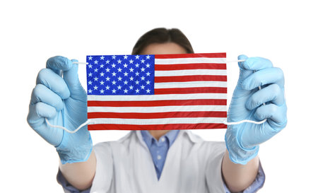 Doctor holding medical mask with USA flag pattern on white background. Dangerous virusの写真素材