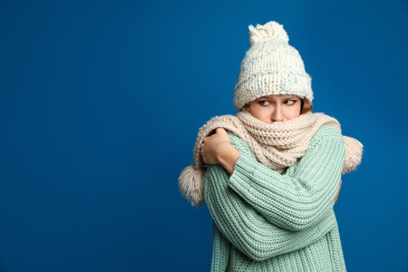 Young woman wearing hat and scarf suffering from fever on blue background, space for text. Cold...