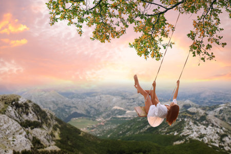 Dream world. Young woman swinging over mountains under sunset skyの写真素材