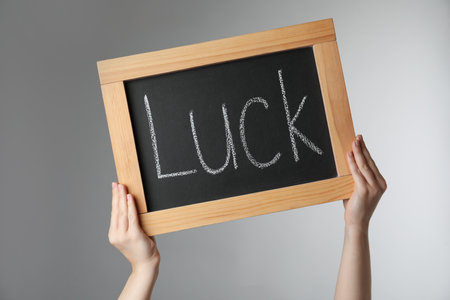 Woman holding blackboard with word LUCK on light gray background, closeup