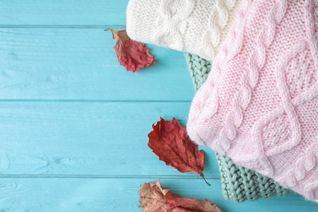 Stack of folded warm sweaters and dry leaves on turquoise wooden background, top view. Space for...