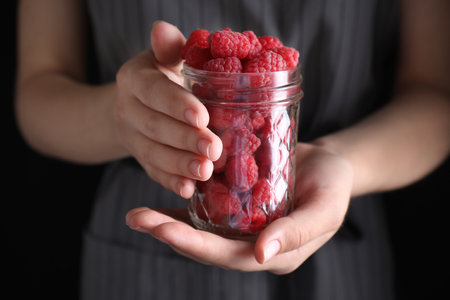 Woman holding glass jar of delicious ripe raspberries on black background, closeupの写真素材