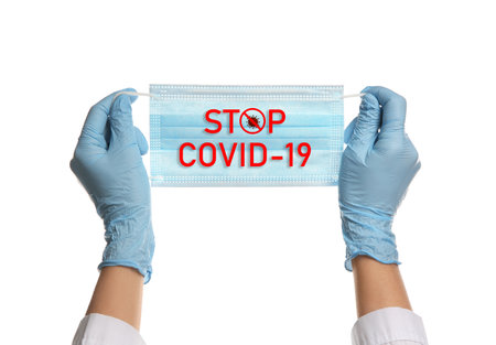 Doctor holding medical mask with text Stop Covid-19 on white background, closeup. Protective measures during pandemicの写真素材