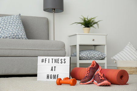 Sport equipment and lightbox with hashtag FITNESS AT HOME on floor indoors. Message to promote self-isolation during COVIDâ19 pandemicの写真素材