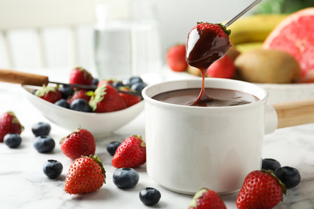 Dipping strawberry into fondue pot with chocolate on white marble tableの写真素材
